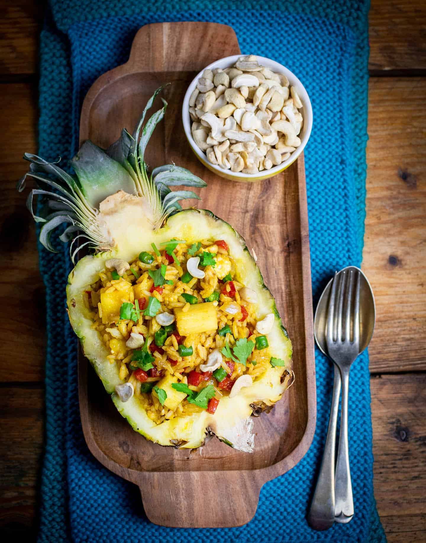A hollowed out half pineapple filled with rice, Cashews in a bowl next to it, with cutlery to the right of the pineapple and a blue cloth underneath it