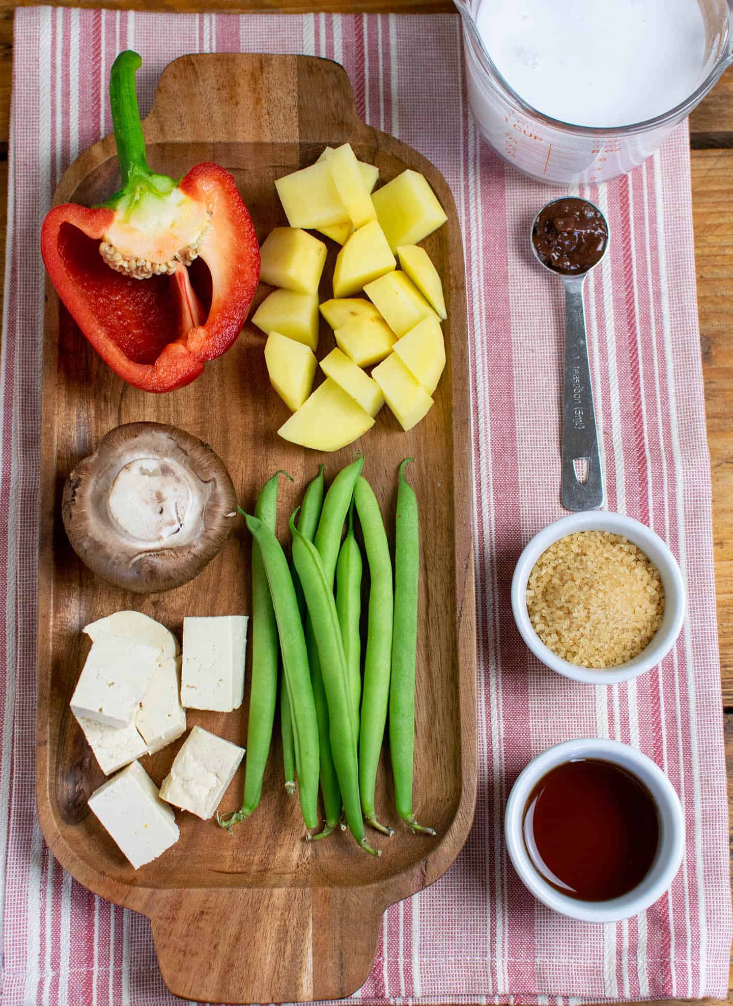 Ingredients for this recipe laid out on a wooden tray. Including green beans, tofu, mushroom, potatoes, red pepper, sugar, tamarind, vegan fish sauce and coconut milk