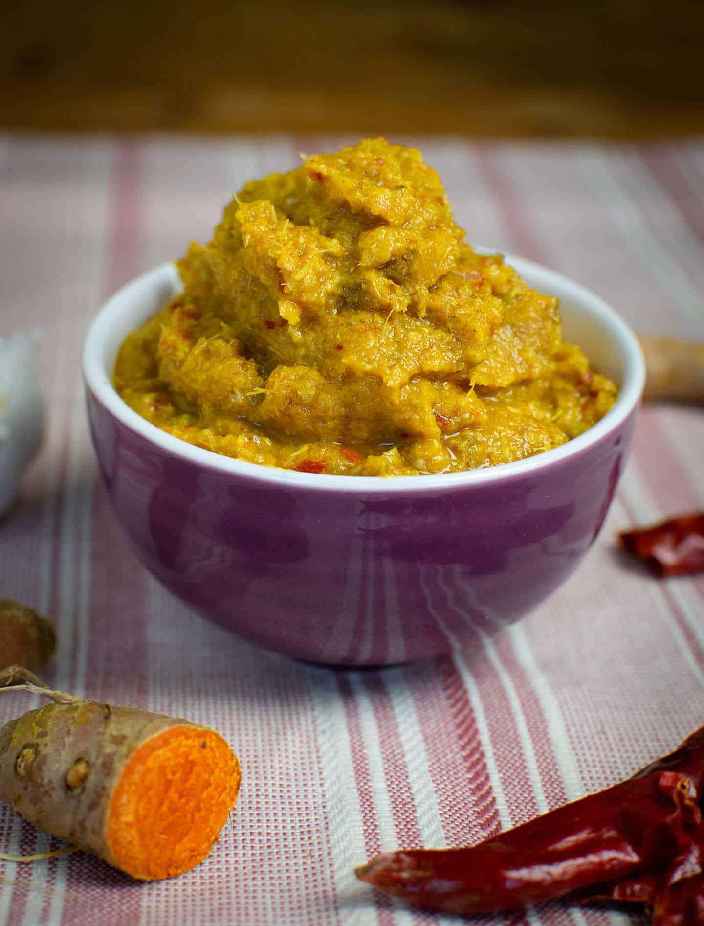 A small purple bowl sat on a stripy pink tea towel, full with homemade yellow curry paste. Turmeric and dried red chillies can be seen around the edges.