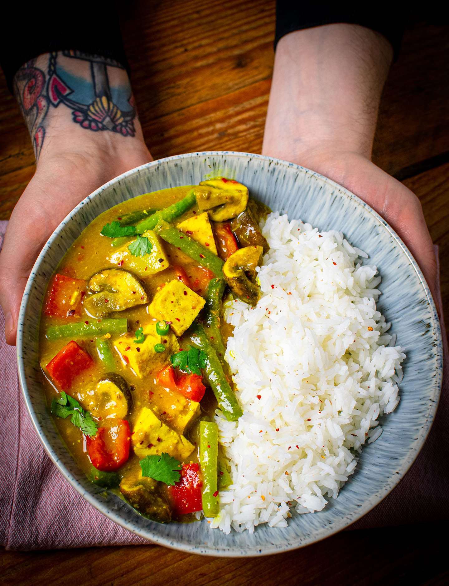 A blue bowl containing the vegan yellow curry. being held up by two hands. Tofu, mushrooms, green beans and red pepper can be seen