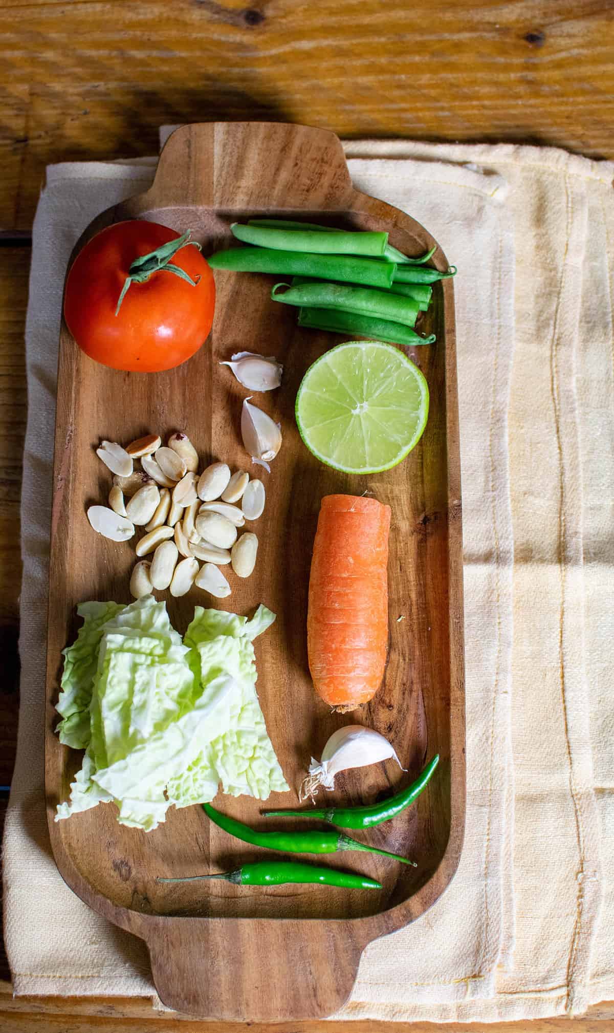 Vegan papaya salad ingredients laid out on a wooden board, showing tomato, beans, lime, garlic, peanuts, carrot, cabbage and green chilli