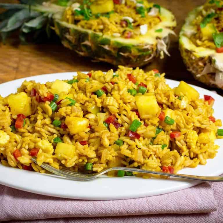 Vegan Pineapple Fried rice served on a white oval plate with a fork on it. There is a pineapple in the background