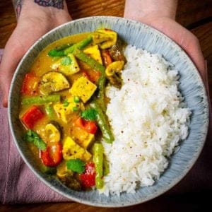 A bowl full of tofu yellow curry being held by Dan. Rice is on one side and curry the other. Dan's wrist tattoo is visible.