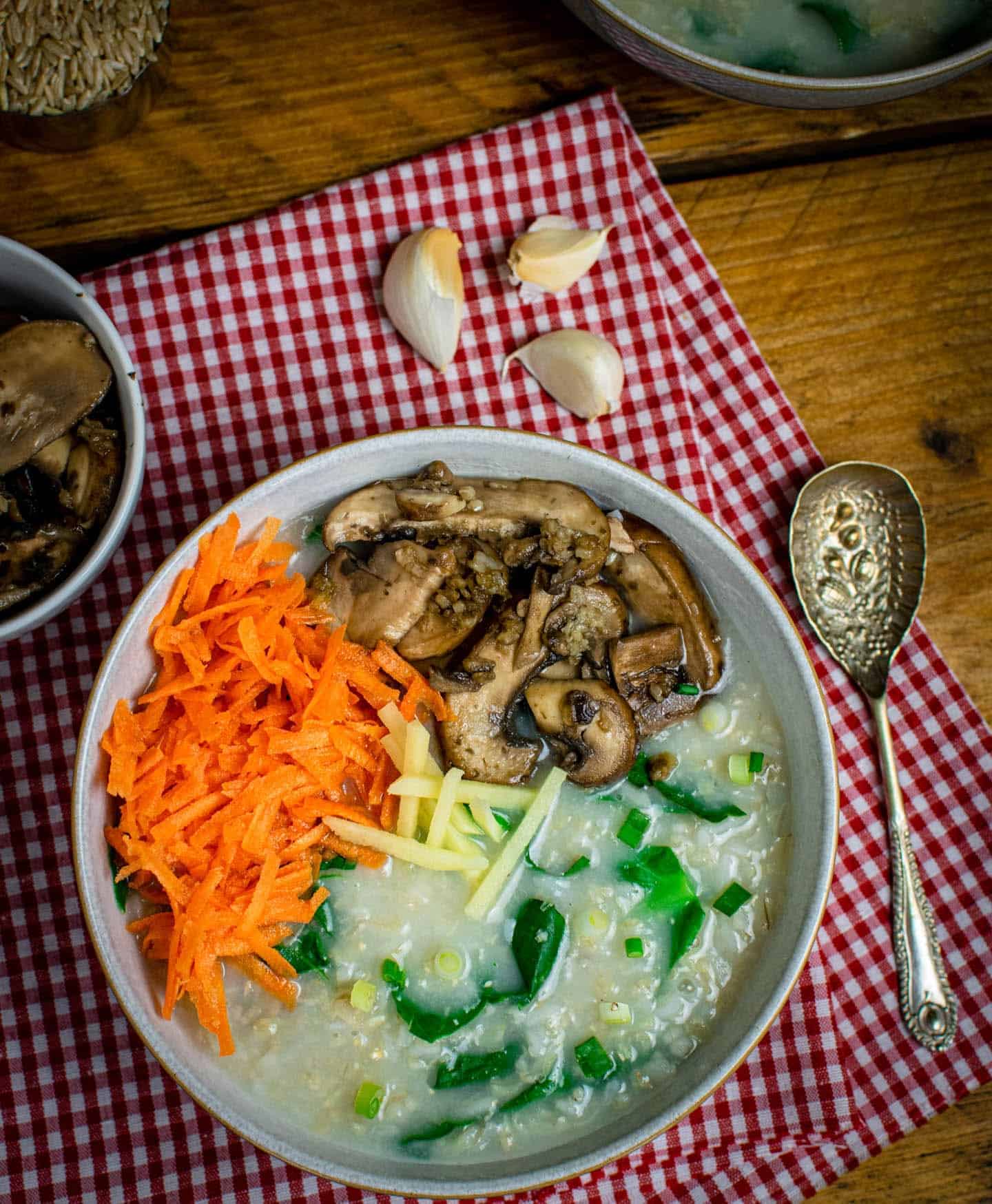 A bowl of vegan congee topped with cooked mushrooms, grated carrot and ginger matchsticks. There's a spoon to the right of the bowl and a red and white gingham cloth underneath it.