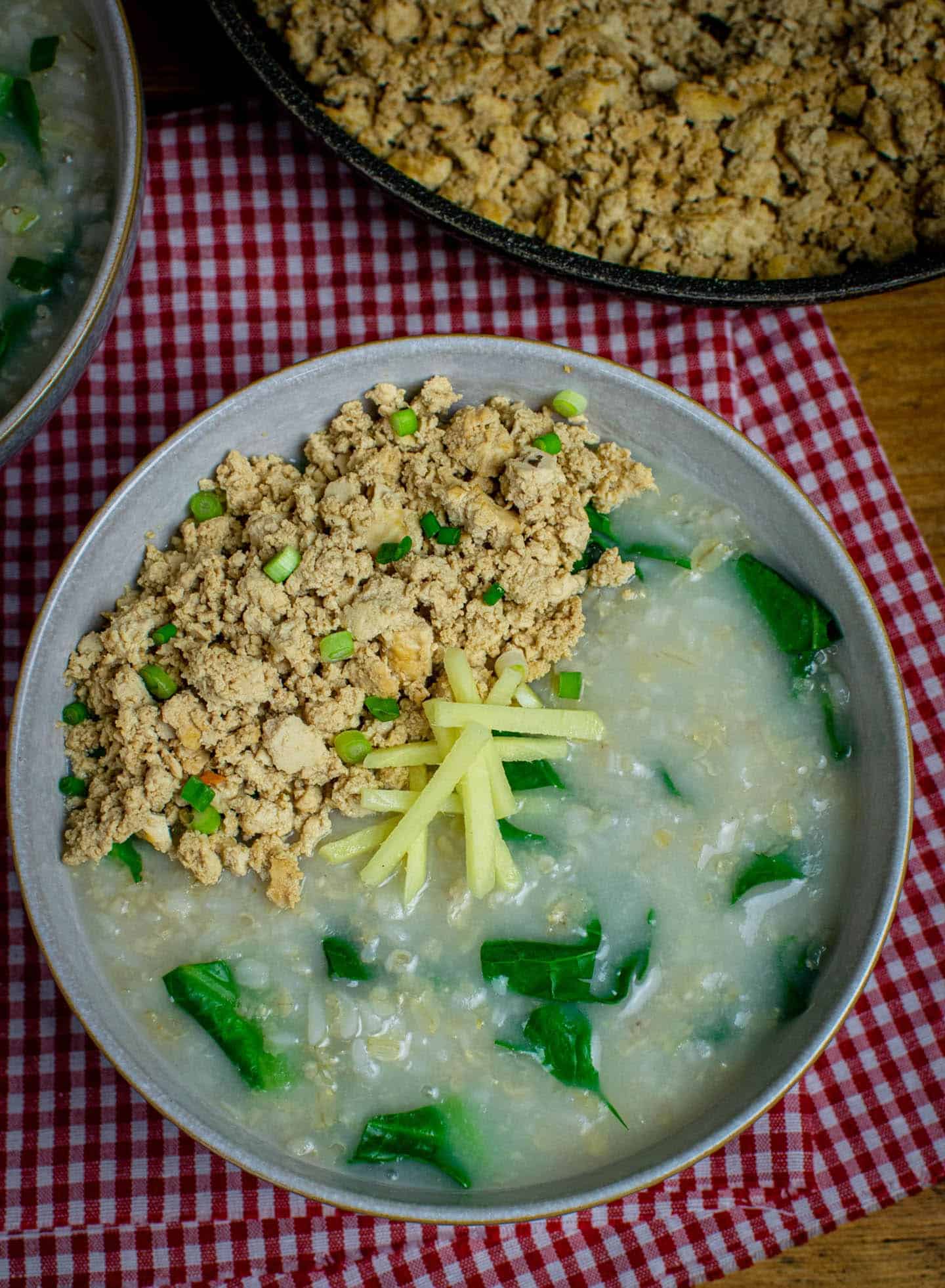 Top down view of a bowl of Thai jok with greens, crumbled tofu, spring onions and thin slices of fresh ginger.