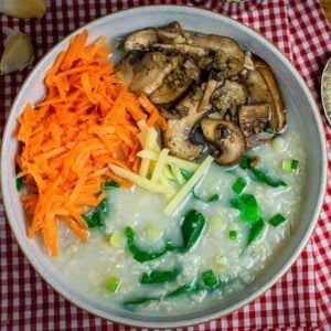 A square cropped photo with a large bowl of porridge as the centre focus. It's topped with grated carrot, fried mushrooms that have garlic on them, fresh slices of ginger and some spring onions.