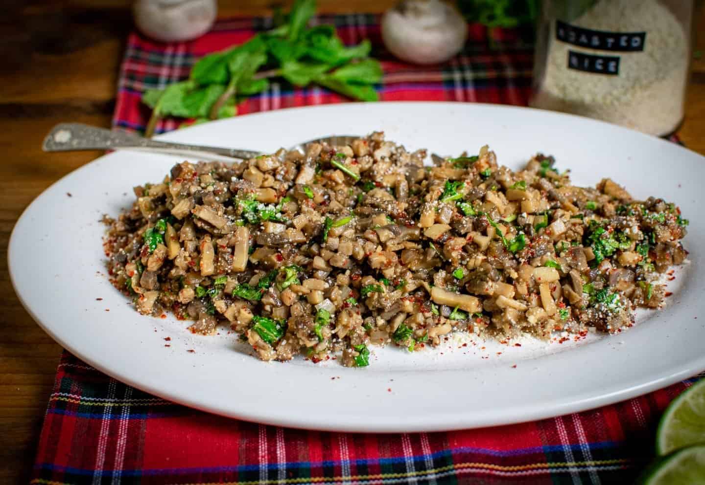 Mushroom larb on a white oval plate with a fork on the plate, a jar of roasted rice, mint and mushrooms in the background