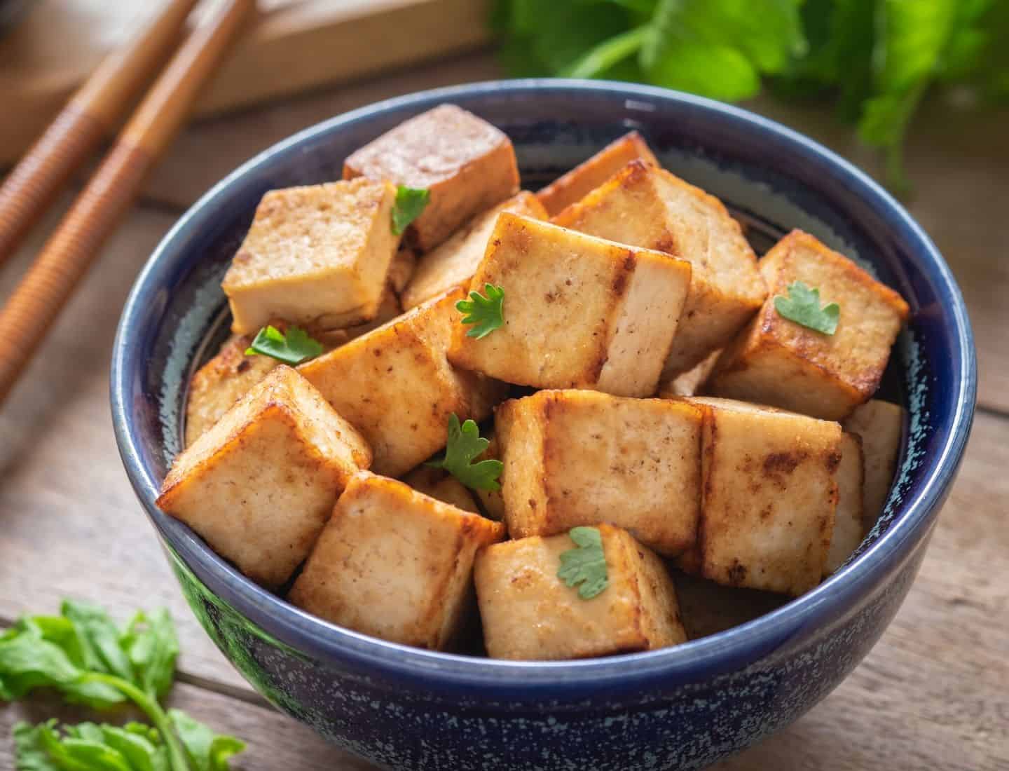 Browned chunks of tofu in a blue bowl with chopsticks in the background