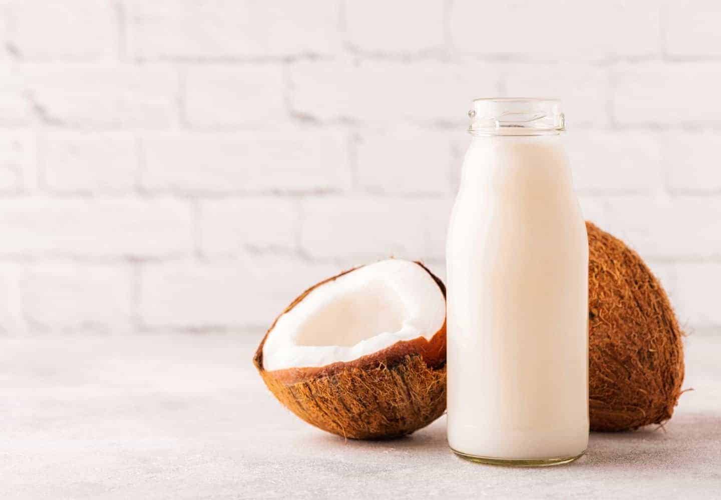 A bottle of coconut milk with two halves of a coconut set against a white brick wall