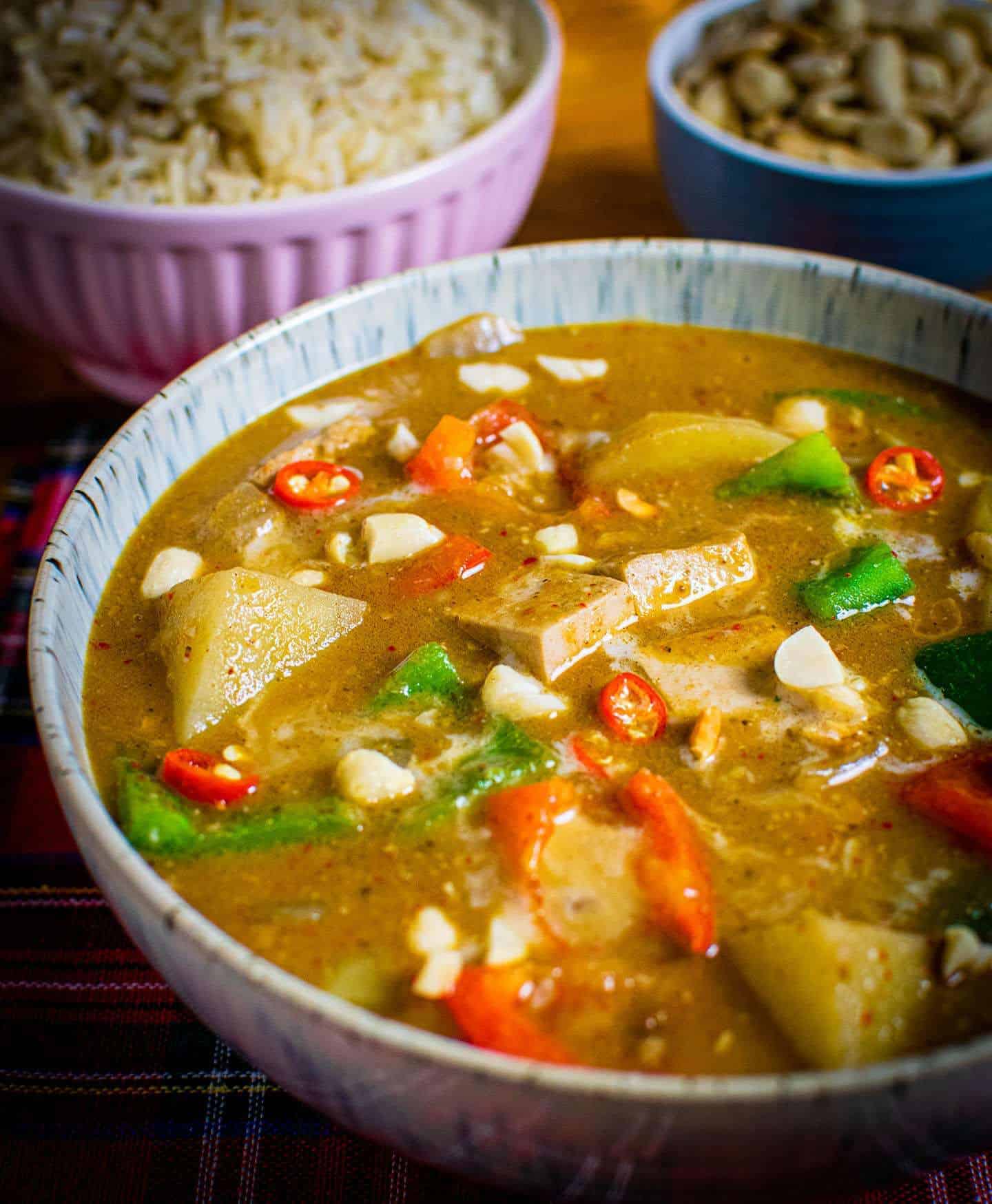 Close up of brown Massaman curry in a bowl, with rice and peanuts in the background. Multiple vegetables are visible in the bowl.