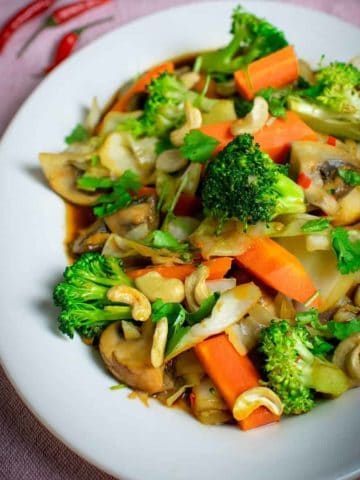 Thai vegetable stir fry on a white oval plate. Broccoli, carrots cabbage and mushrooms and cashews can be seen. Set on a pink background.