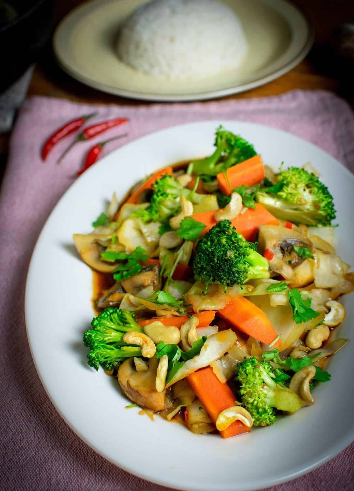 Veggies on a white oval plate in a Thai stir fry sauce with chillies in the background and a dome of rice on a yellow plate is visible in the background too