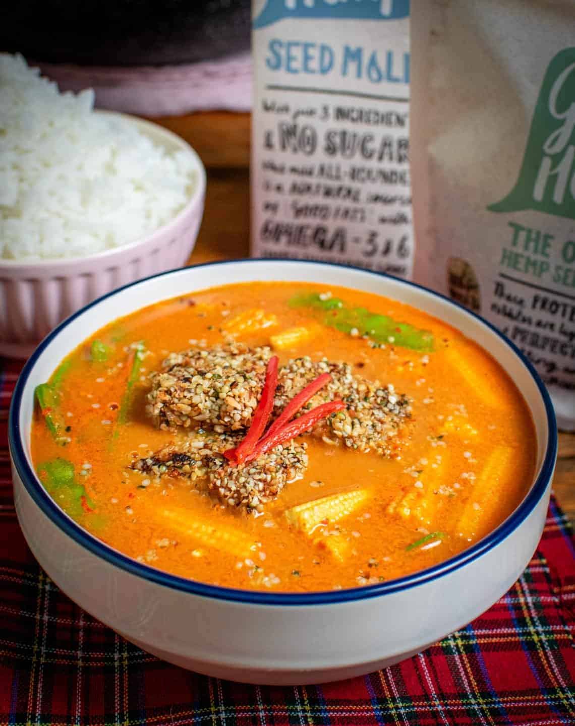 A white bowl full of Thai red curry with tofu and chilli on top, there's products in the background as well as a bowl of rice