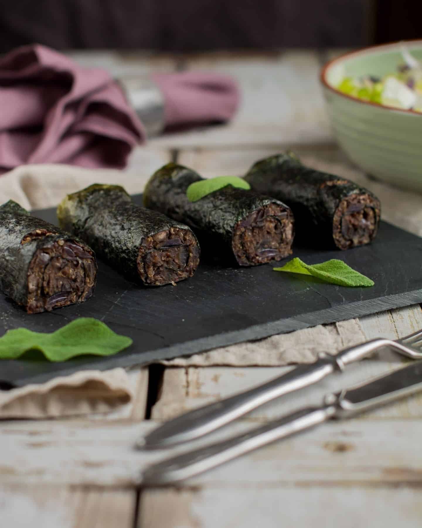 4 nori rolls filled with purple rice on a grey slate with a tea towel in the background