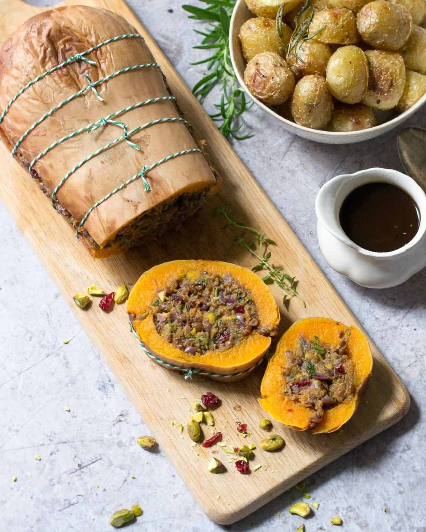 Stuffed butternut squash on a chopping board with two slices cut off, a gravy just and a bowl of cooked potatoes