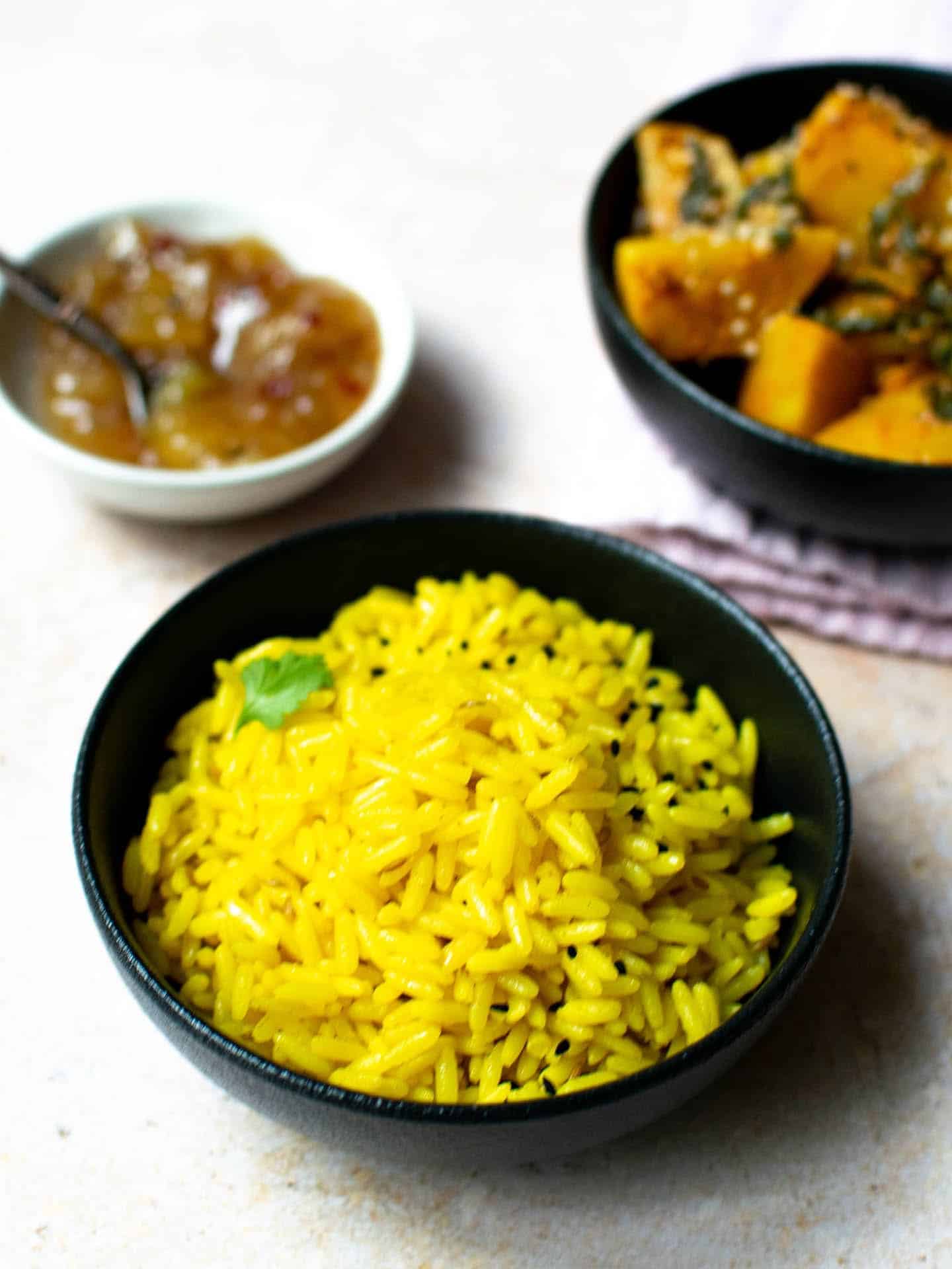 A close-up image of a serving of golden pilau rice in a black bowl. In the background is a potato dish and a small bowl of chutney. The rice in the foreground is sprinkled with some nigella seeds and a bit of fresh coriander.