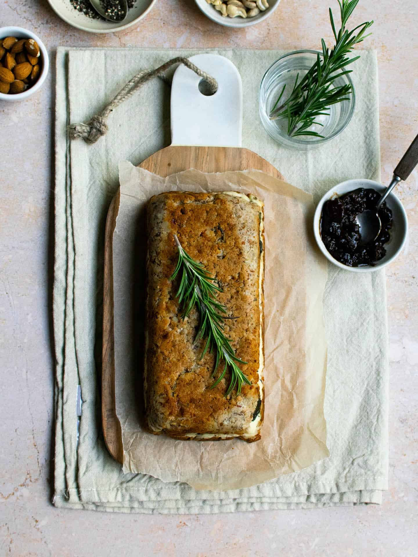 A top down photo of the cashew nut roast, sitting on top of a chopping board, which is on top of a stone coloured tea towel. There are a few pots on show round the board, one has rosemary in, another has a chutney or sauce in it. The cashew roast is a golden brown colour and is in a loaf shape.