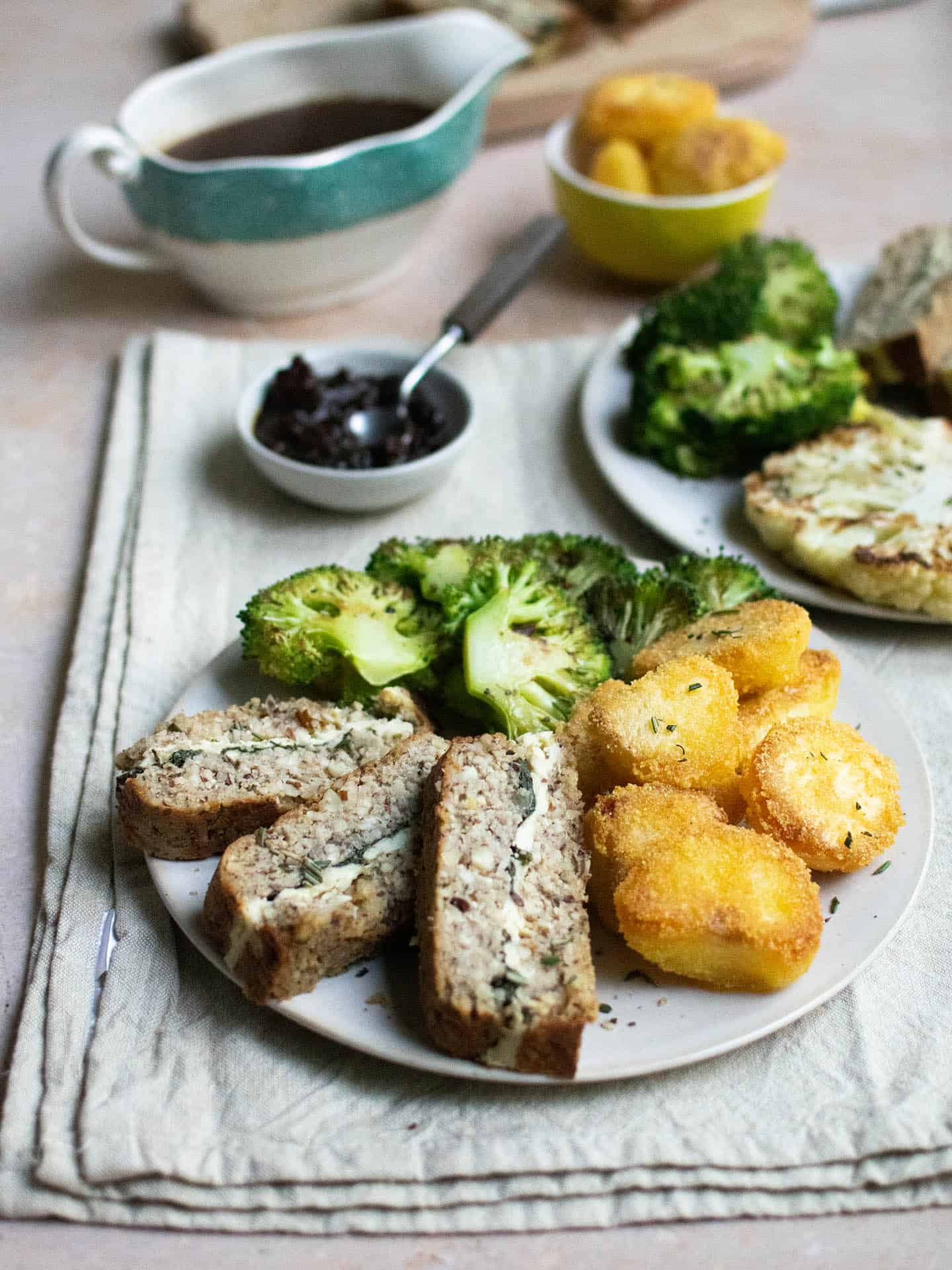 A photo with a plate filled with food at the forefront of it. The plate has slices of cashew nut roast, roast potatoes and broccoli on it. In the background is a jug of gravy, a small bowl of roast potatoes and to the side is a second plate which has a roasted cauliflower steak on, as well as the nut roast and broccoli.