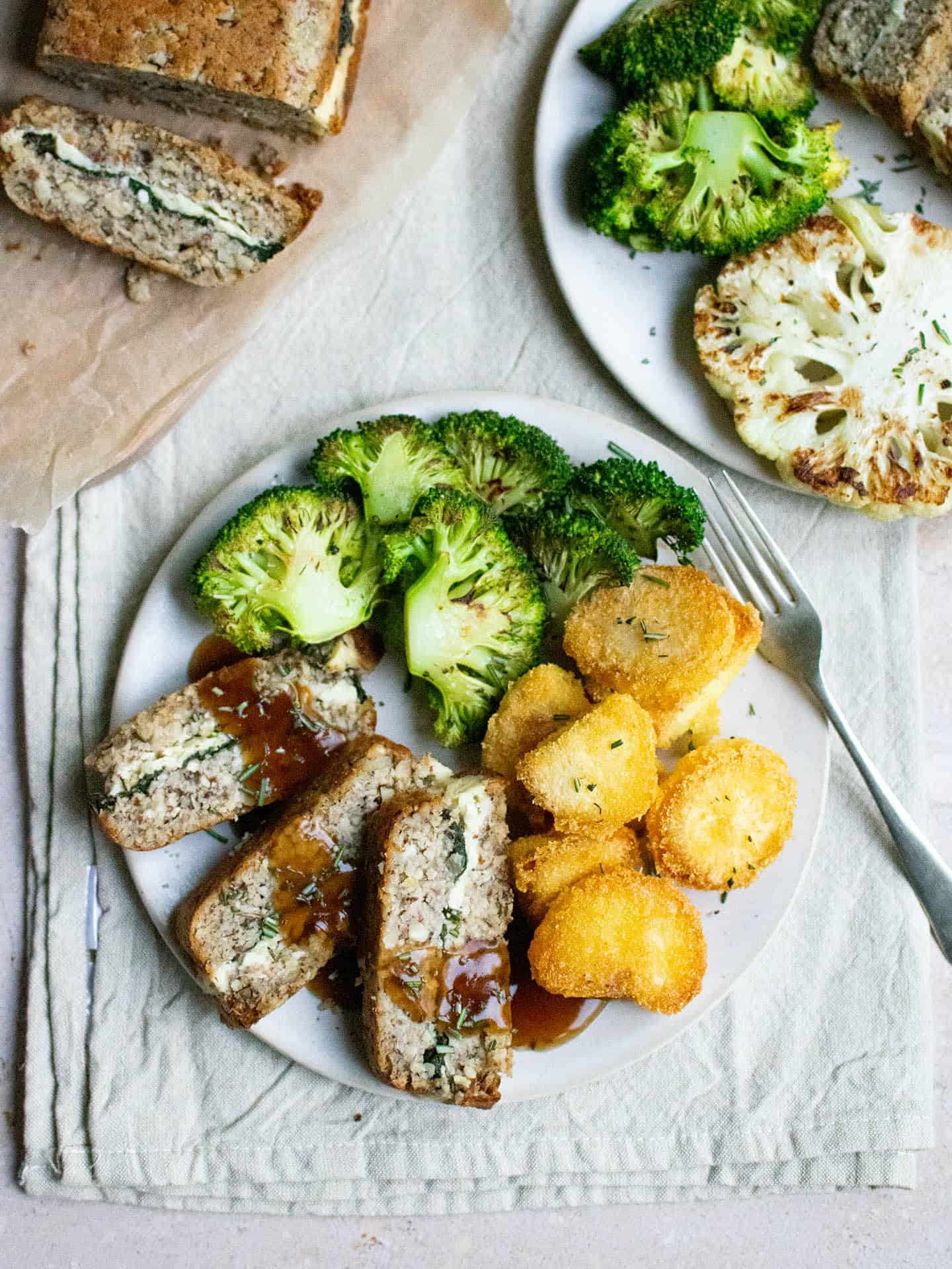 A photo with two plates in it. The plate at the front has nut roast, roast potatoes and broccoli on it. There is a drizzle of gravy over the nut roast. In the top left corner is the whole cashew nut roast with a slice cut off and laying face up. The second plate in the right of the photo has a grilled cauliflower steak on it as well as some broccoli.