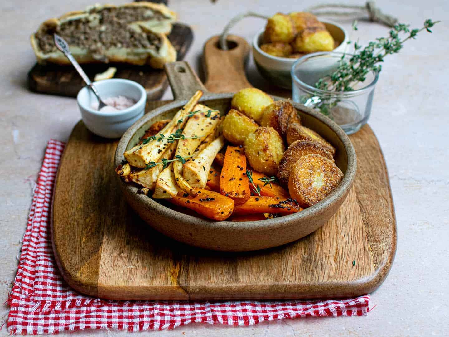 Vegetables in a circular serving dish with salt, potatoes and mushrooms wellington in the background. The vegetables are sat on a wooden chopping board 