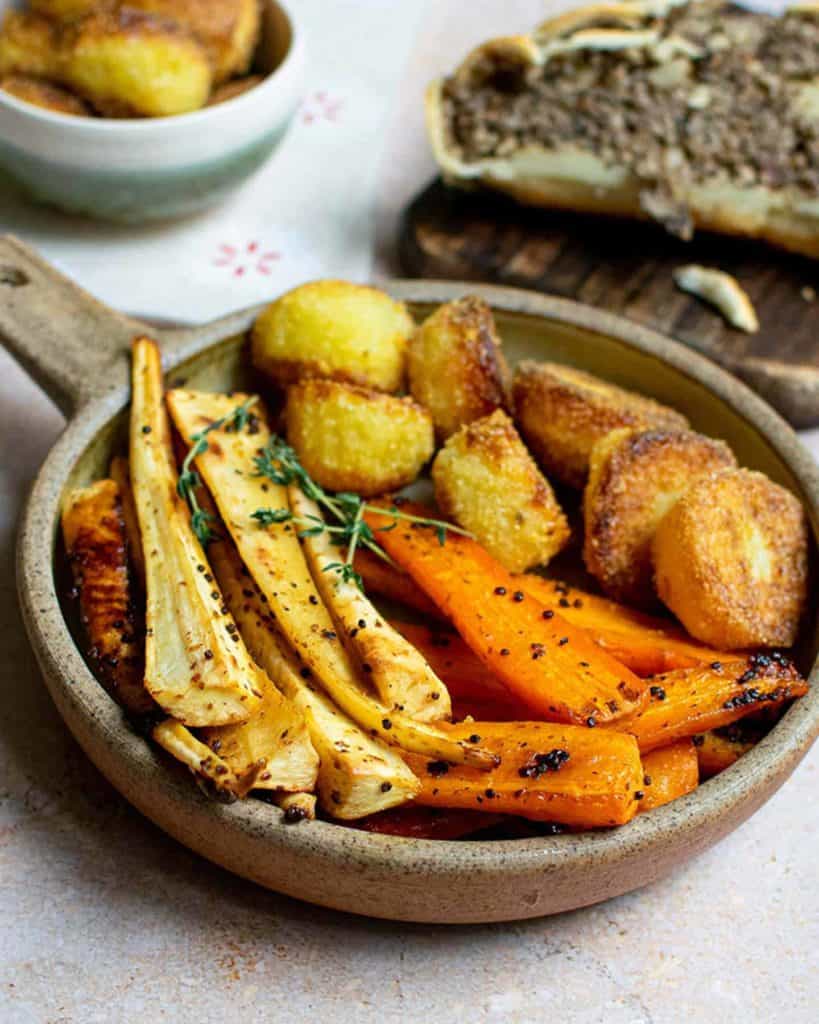 Maple roasted carrots and parsnips in a round dish with roast potatoes and other dishes in the background