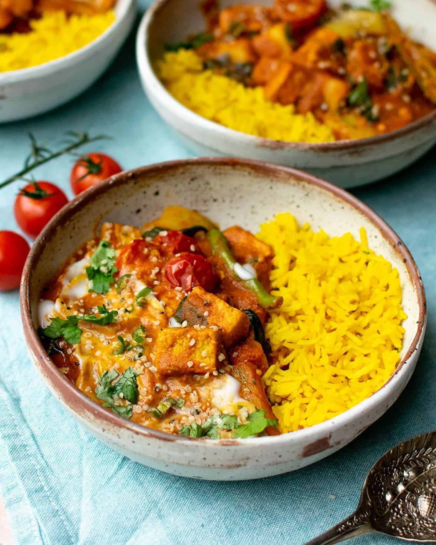 Veg jalfrezi in a speckled bowl, red curry with quorn pieces can be seen and there's yellow pilau rice to the right of the bowl. In the background there is more bowls of curry and cherry tomatoes