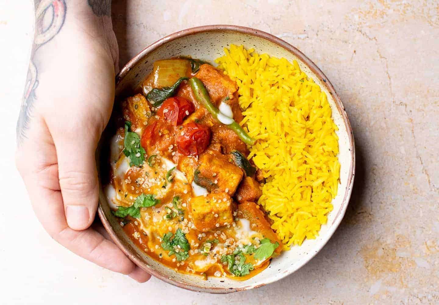 A bowl of veg jalfrezi being held with one hand, and tattoos can be seen on the top of the hand. Coriander, pilau rice, tomatoes, onion and quorn can be seen in the bowl