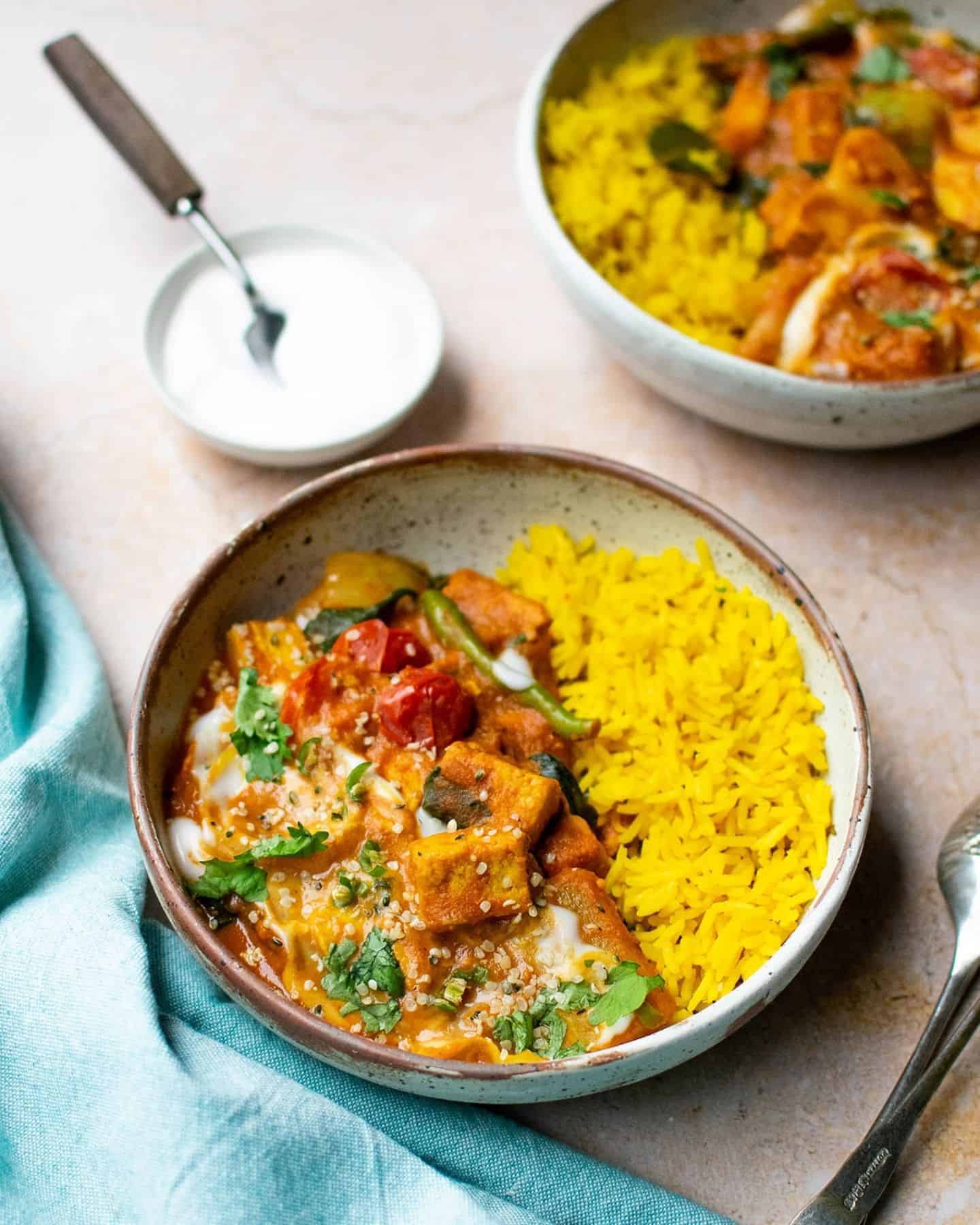 Curry and pilau rice in a speckled bowl with a blue cloth to the left, another bowl of curry in the background to the right and a small dish of yoghurt with a spoon in it to the left in the background