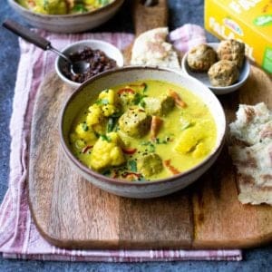Falafel curry in a bowl on a wooden board with chutney and more falafel behind it with naan to the right side.