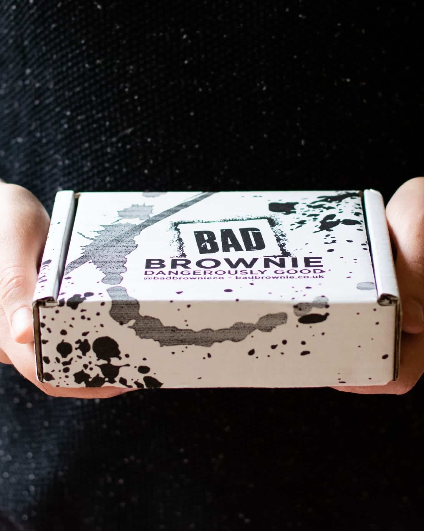 A set of hands holding a box. The box says Bad Brownie – Dangerously Good. It has a black and grey paint splatter effect as the design.