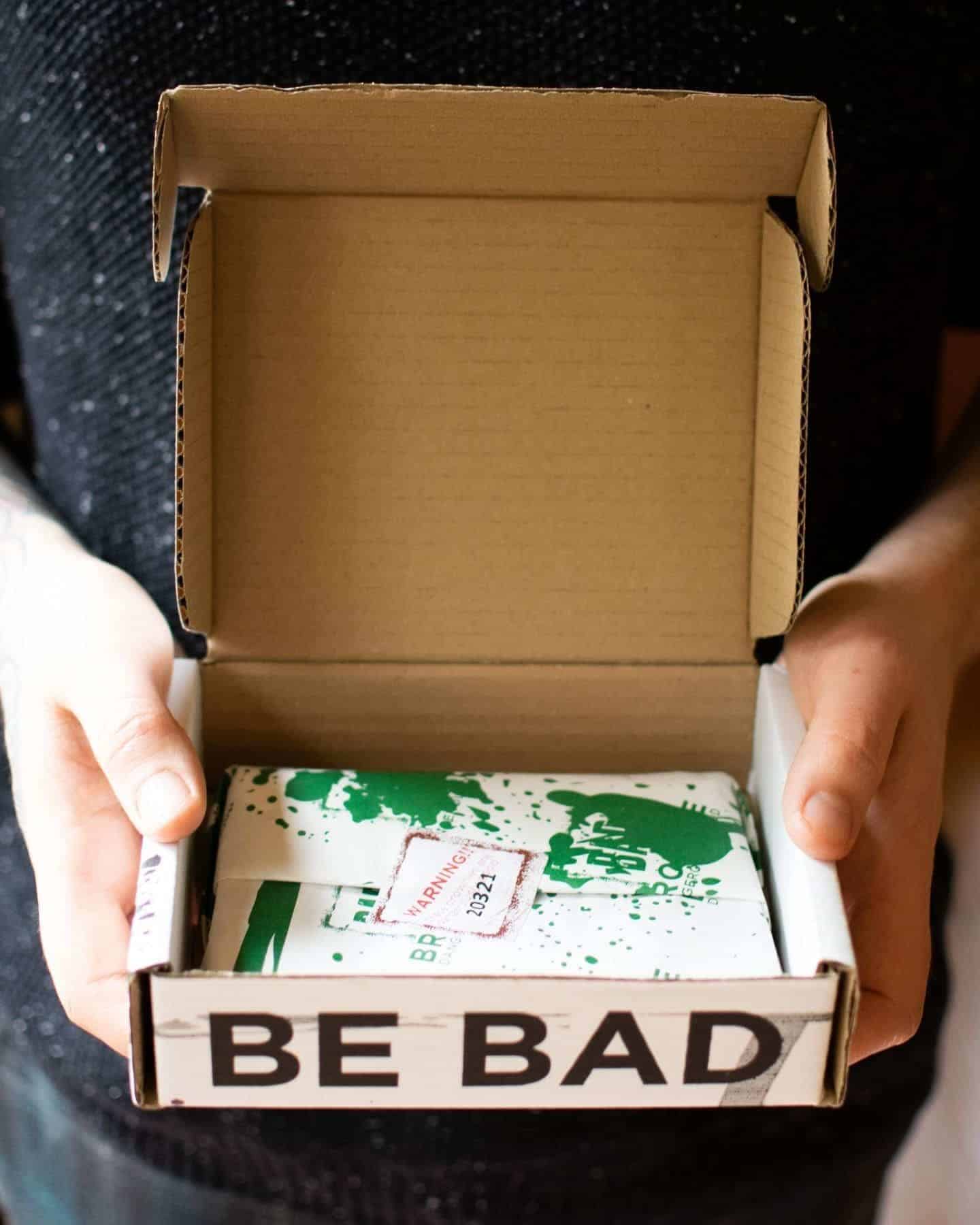 The box has been opened revealing a message "Be Bad". Inside is some green paint splattered paper wrapped around the brownies.