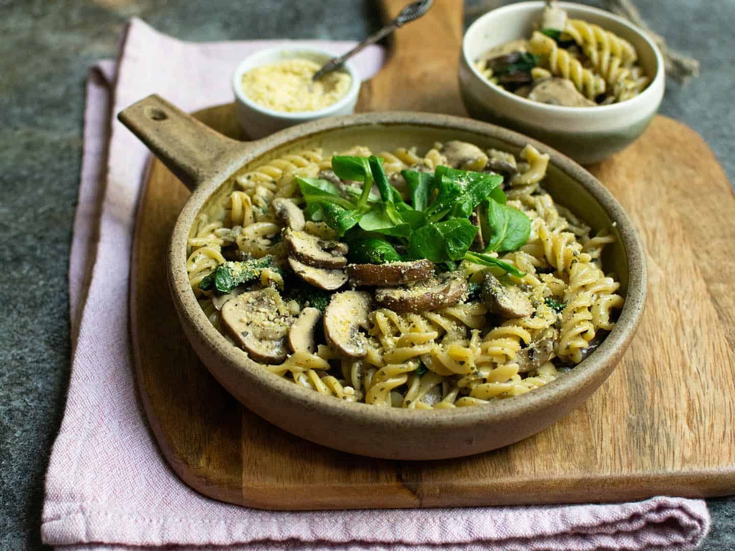 A bowl of vegan mushroom pasta presented on a chopping board with a small bowl of nut cheese.