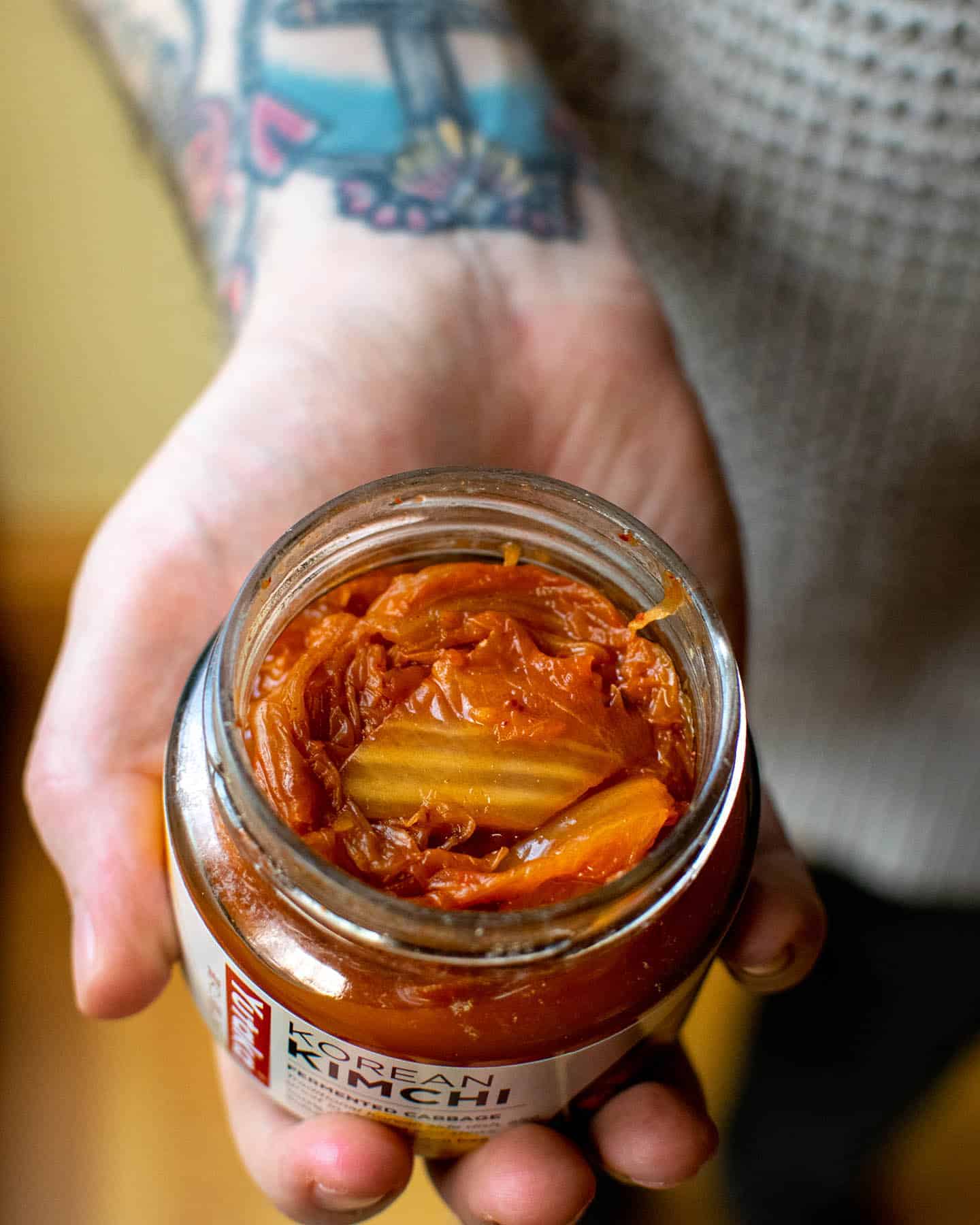 An open jar of kimchi being held by a man's hand with tattoos visible on his wrist