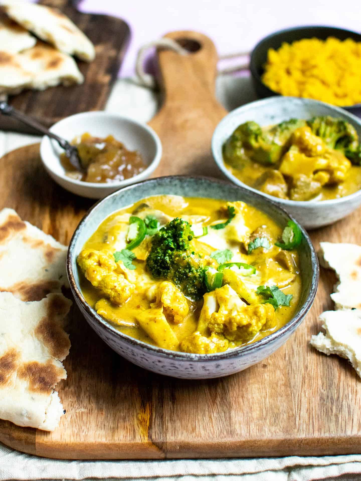 A wooden board showing a bowl of curry in the foreground, and numerous other bowls in the background, including chutney, rice and naan bread on another board