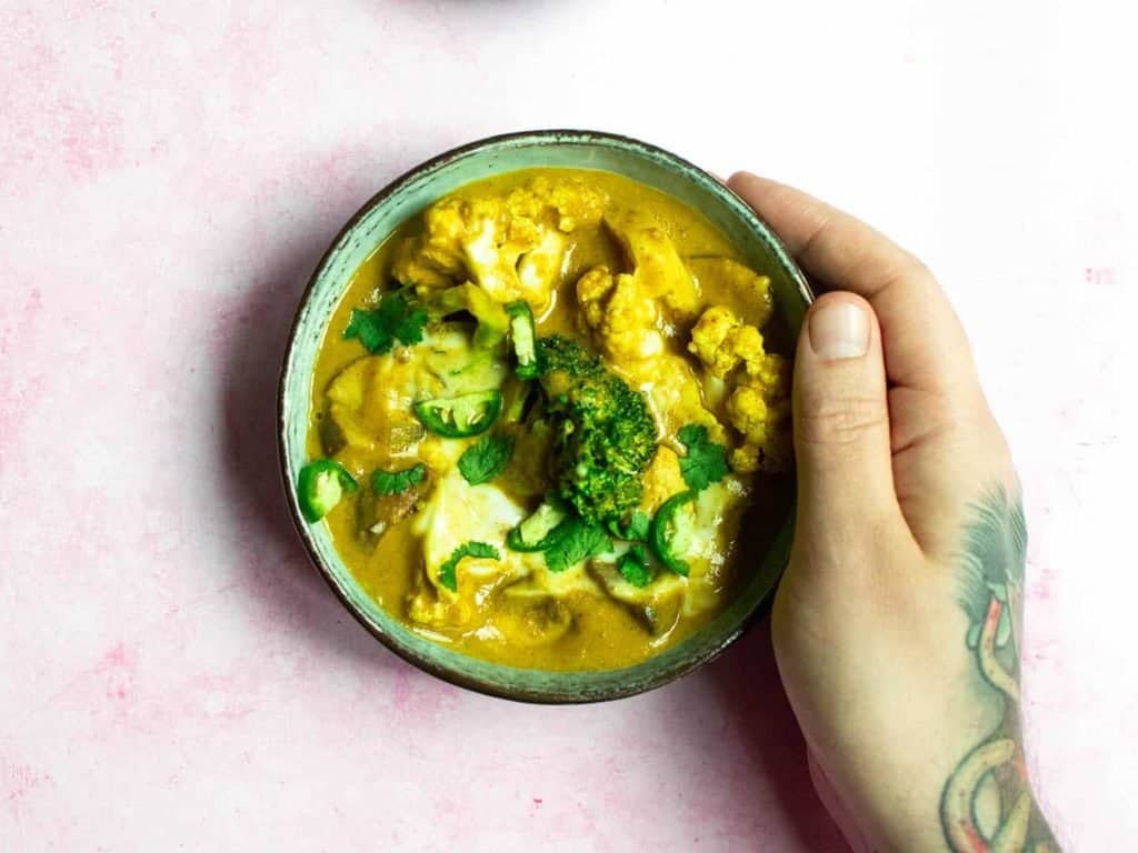 A top down view of a bowl of curry being held by a man's hand, on a pink background