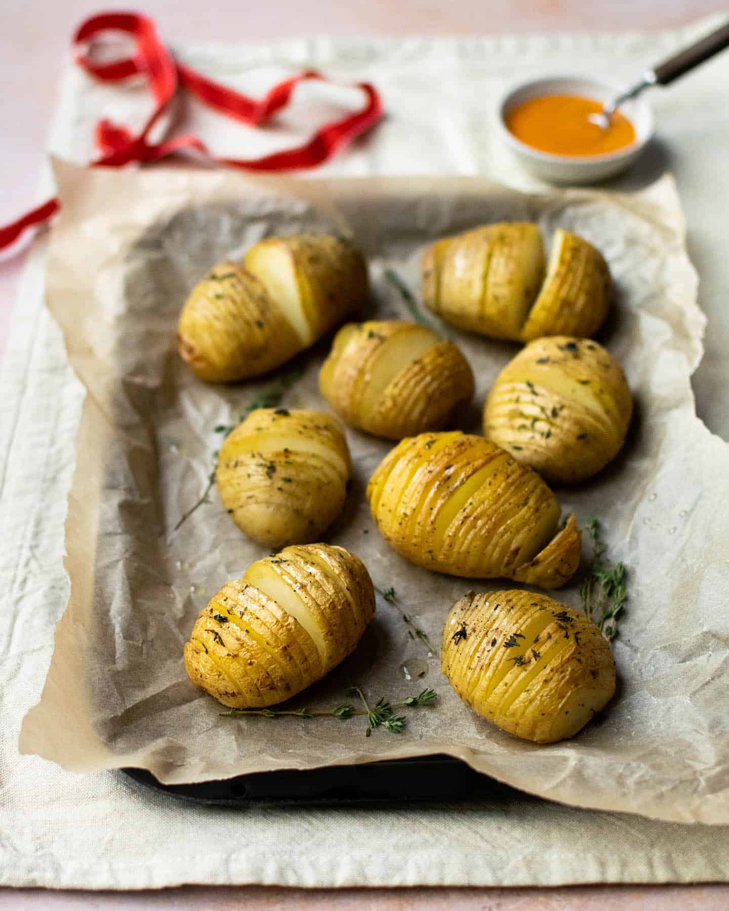 Vegan hasselback potatoes on a tray on greaseproof paper with a ribbon in the background as well as a small dish of sauce with a spoon in it