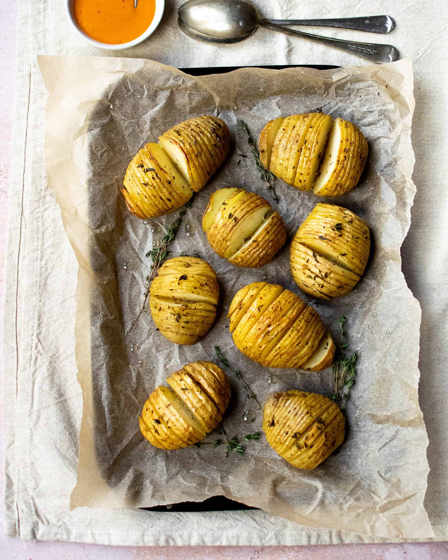 Top down view of herby hasselback potatoes on a tray with a spoon above them, next to a small dish of orange sauce