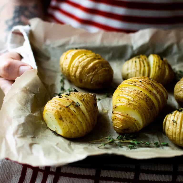 A man wearing a red and white striped t-shirt holding a baking tray of vegan hasselback potatoes, with greaseproof paper on the tray