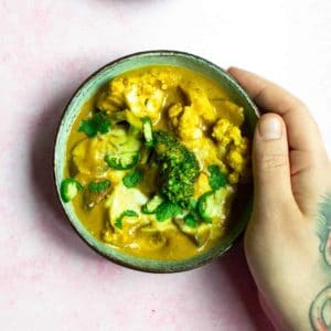 A tattooed hand resting his hand on the side of a bowl that contains peanut butter curry, where broccoli and cauliflower are in a brown sauce