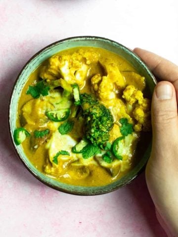 A tattooed hand resting his hand on the side of a bowl that contains peanut butter curry, where broccoli and cauliflower are in a brown sauce