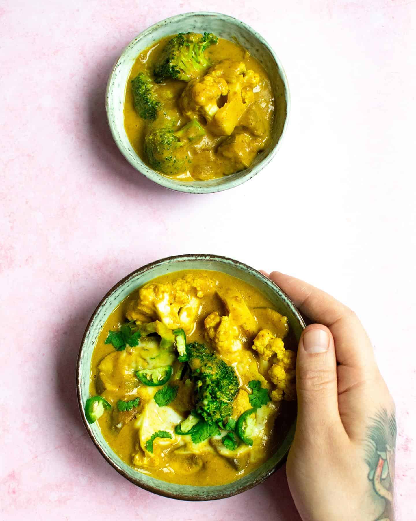 Two bowls of peanut butter curry, one above the other with a man's hand holding one of them