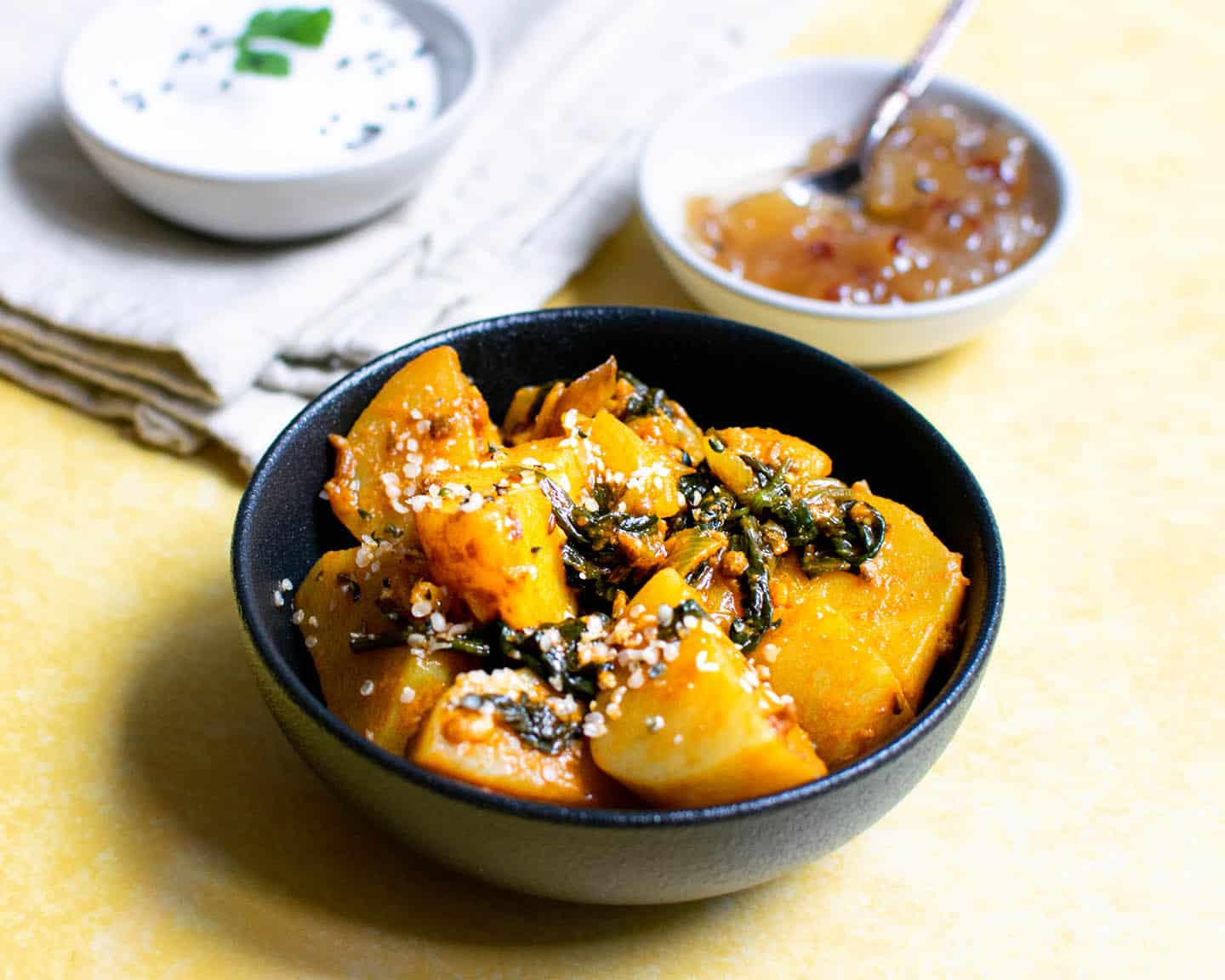 Indian potatoes with spinach in a black bowl on a yellow background with other small pots in the background