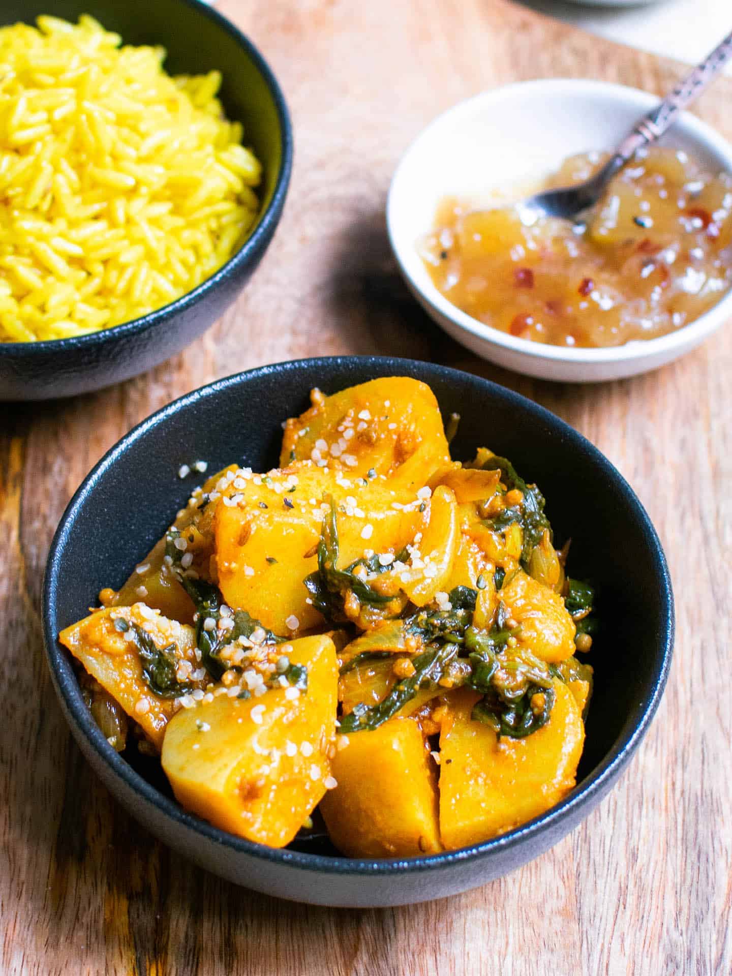 Saag Aloo in a black pot on a chopping board with rice and chutney in the background in separate dishes