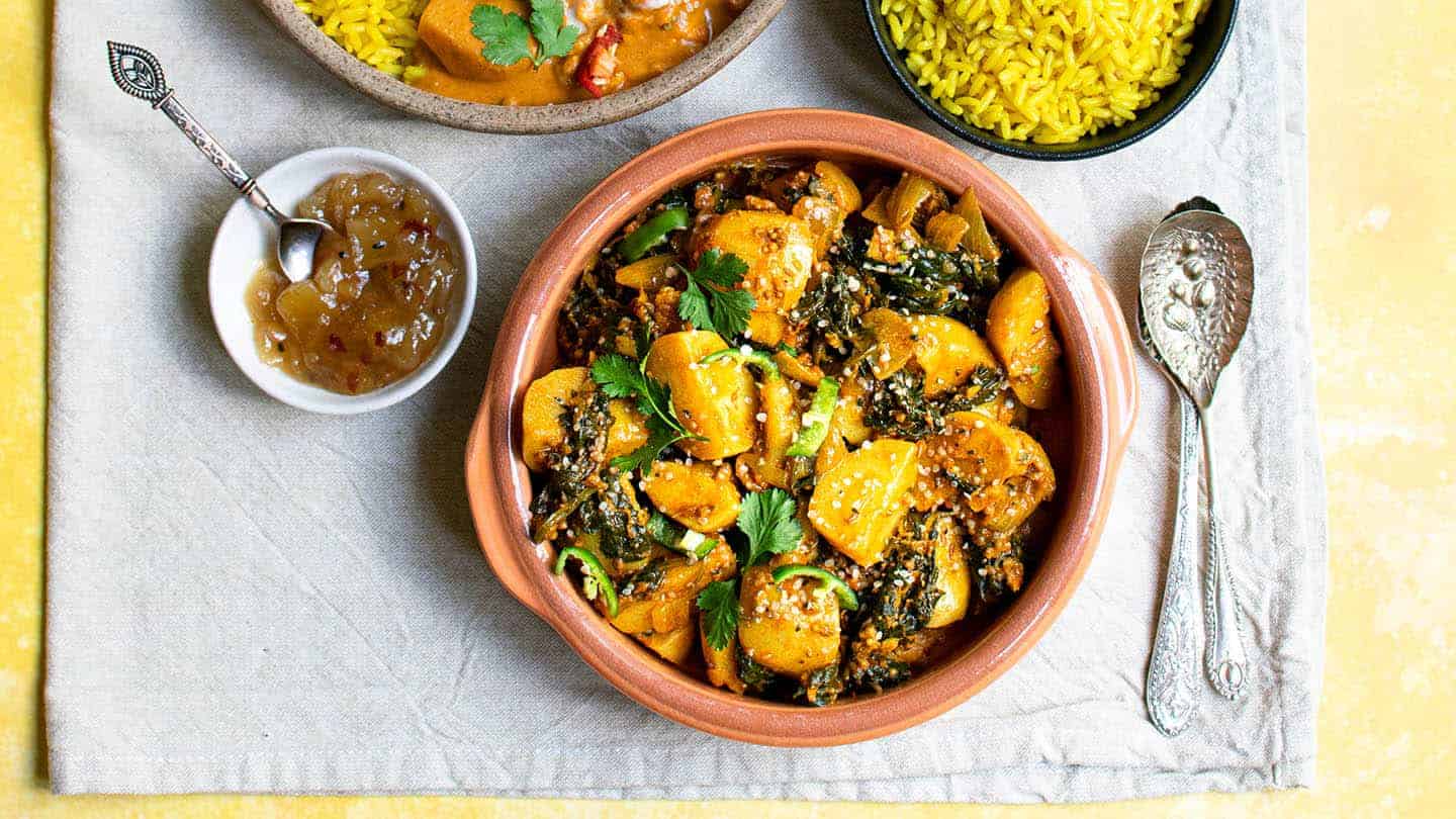 Saag Aloo just off centre to the right with chutney in a small bowl to the left, spoons to the right and rice and curry above it.