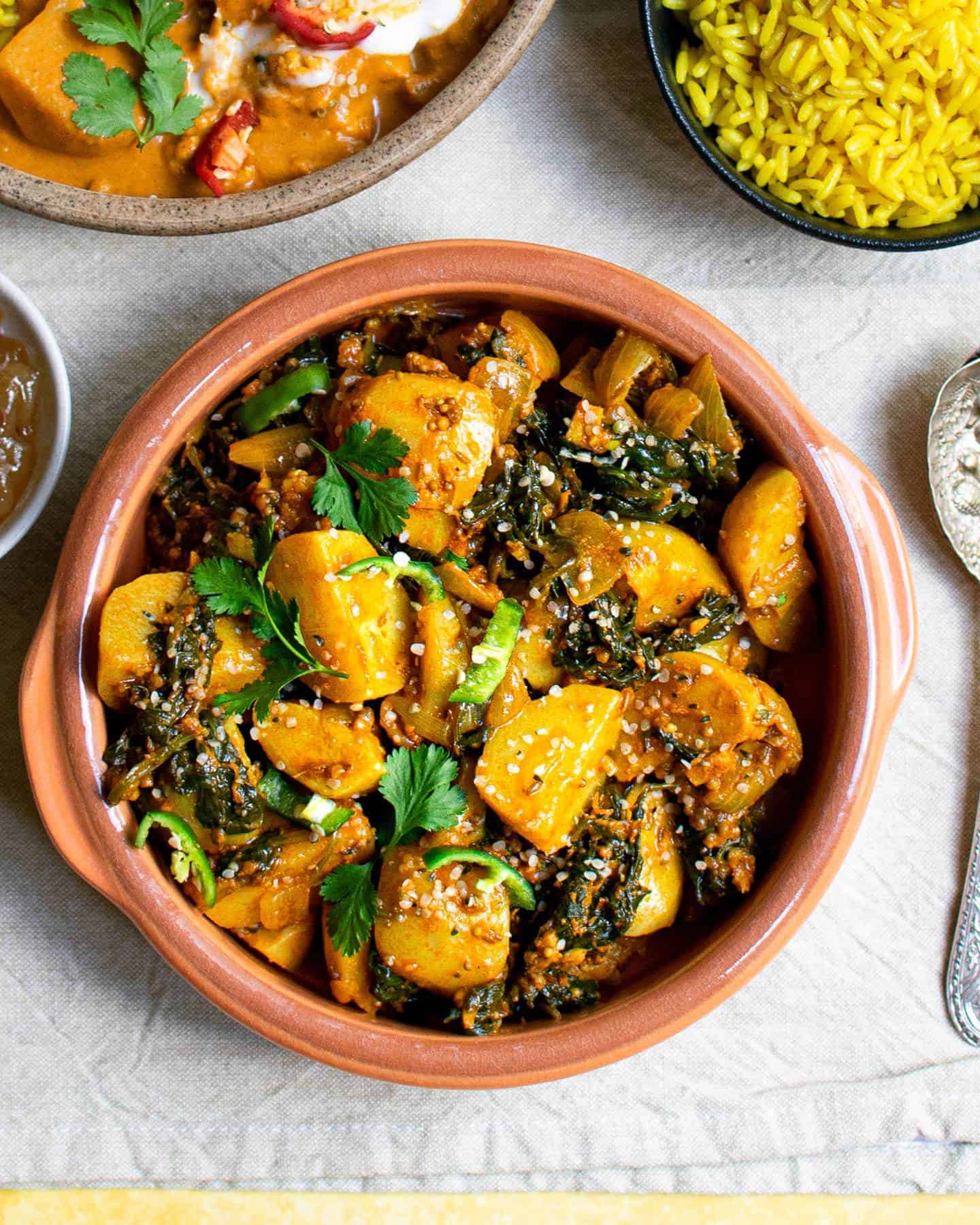 vegan Saag Aloo in a brown bowl with small indented handles, spoons to the right and other curries around it in bowls
