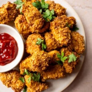 Cornflake coated vegan chicken nuggets piled onto a plate with tomato ketchup.