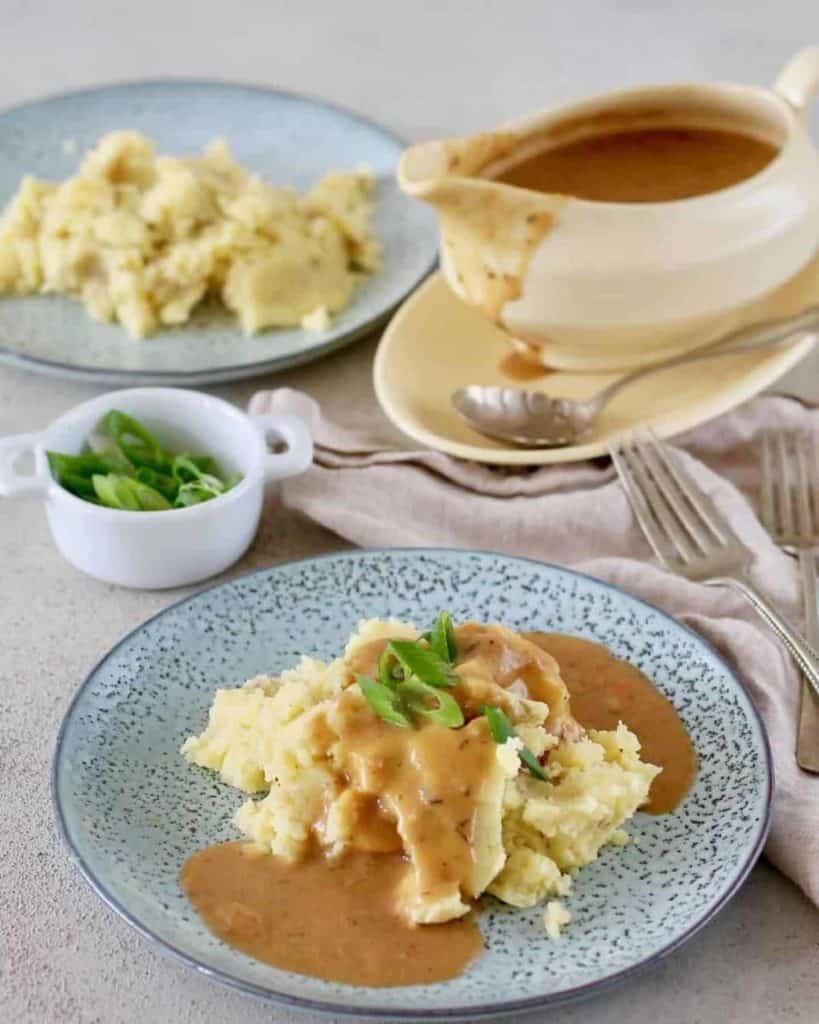 Vegan gravy poured on mash potatoes with other blue plates and a bowl of spring onions in the background