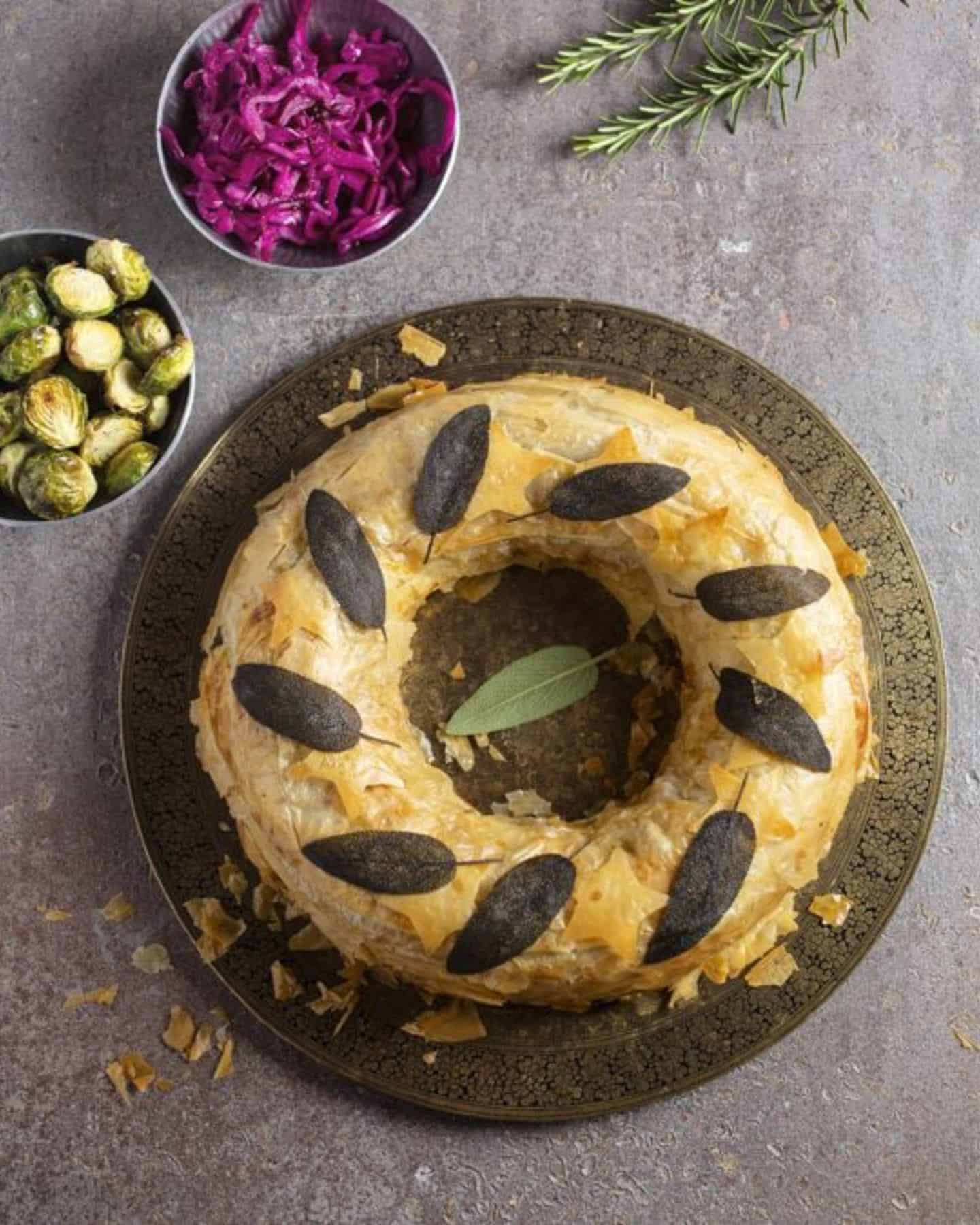 A pastry wreath with vegan roast dinner dishes in the background, showing sprouts and cabbage