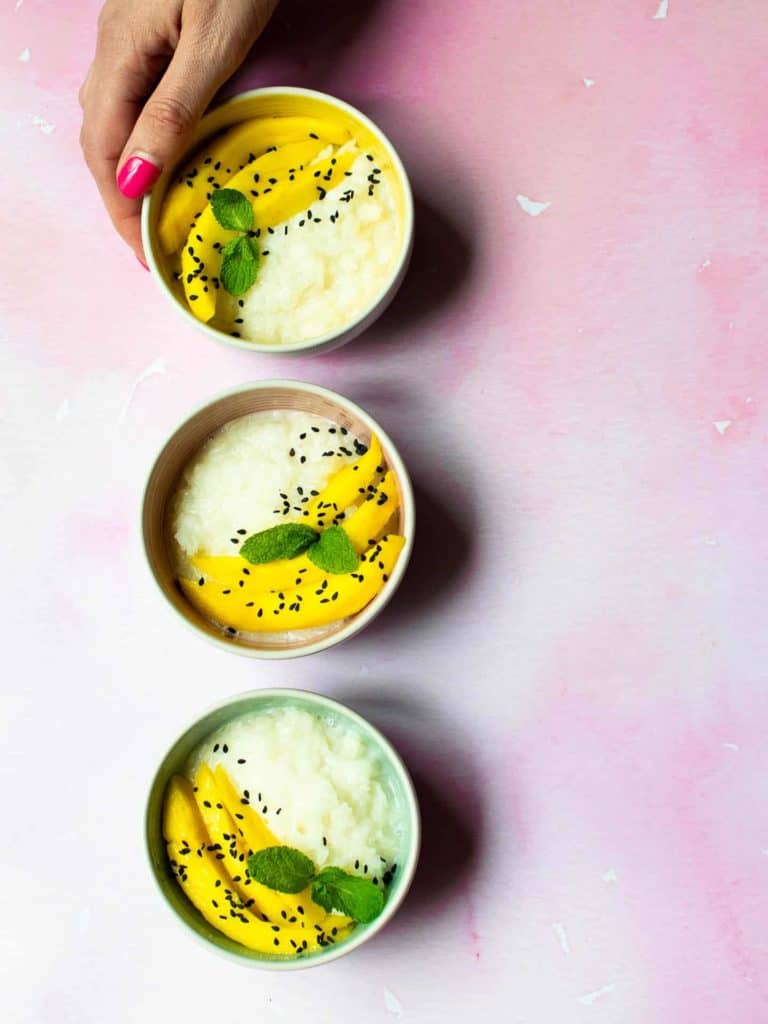 Three bowls in a line, on top of a pink work top. The bowls have sticky coconut rice, slices of mango and fresh mint in them.