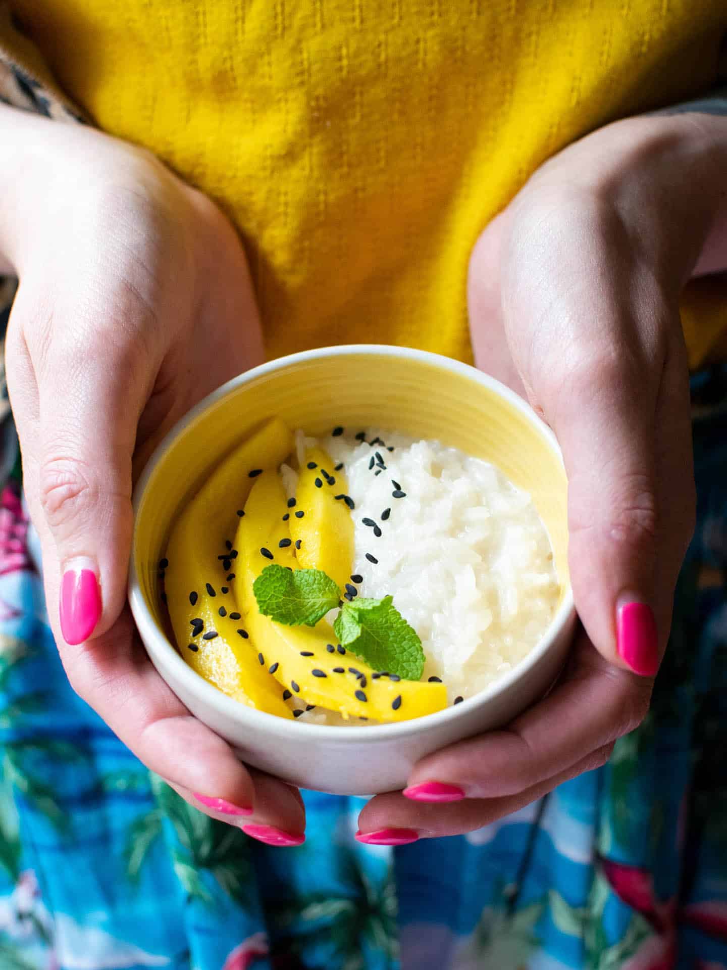 A pair of hands with painted pink nails, holding a bowl of mango and sticky coconut rice.